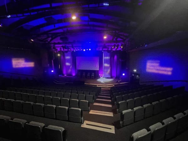 Banked auditorium seating facing a stage and screens under blue lighting at a corporate conference